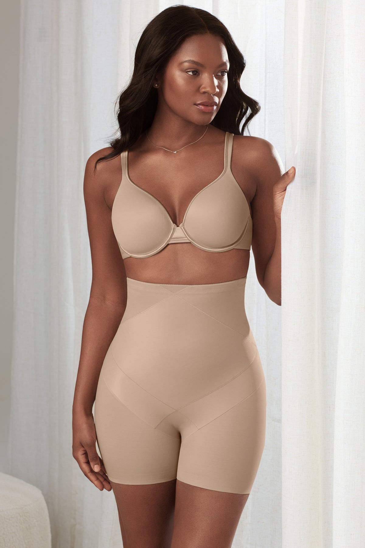 a woman looking out a curtain while wearing a nude high waisted body shaper short shapewear