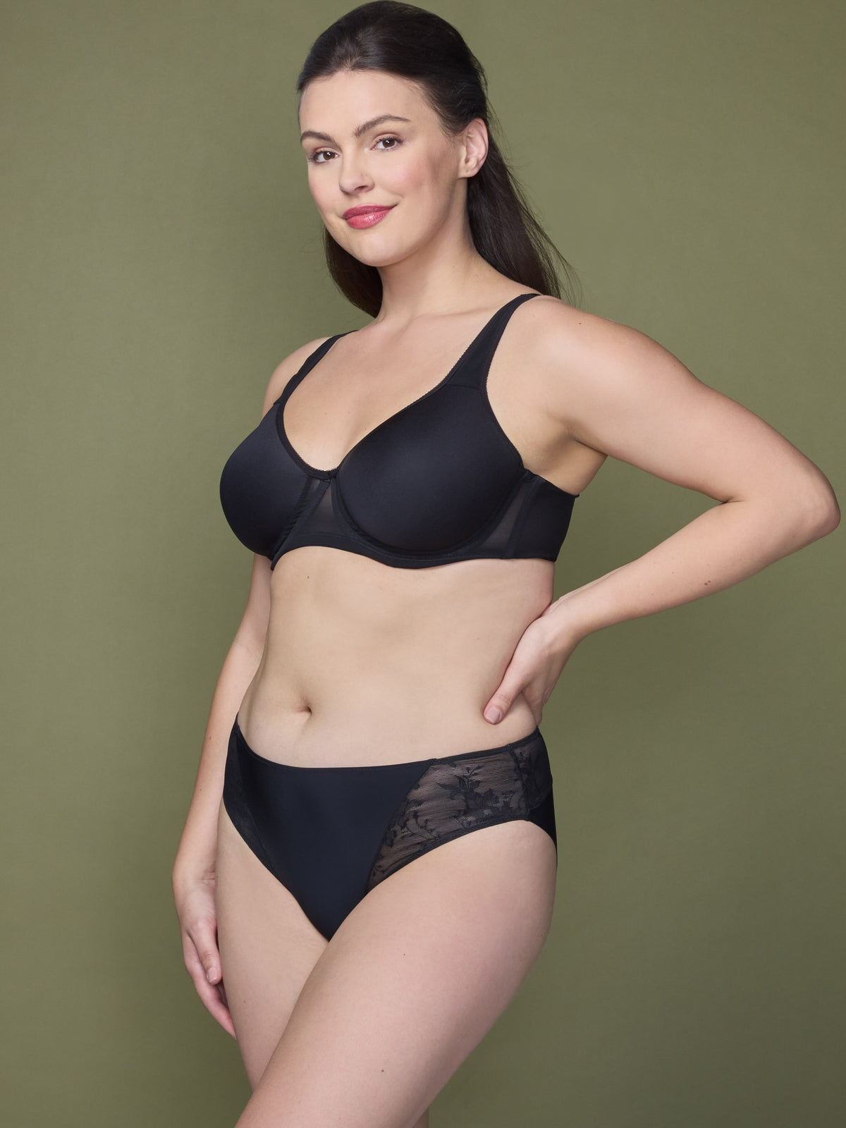 a woman standing against an olive backdrop with her hand on her hip wearing a black lace hipster panty all-groups
