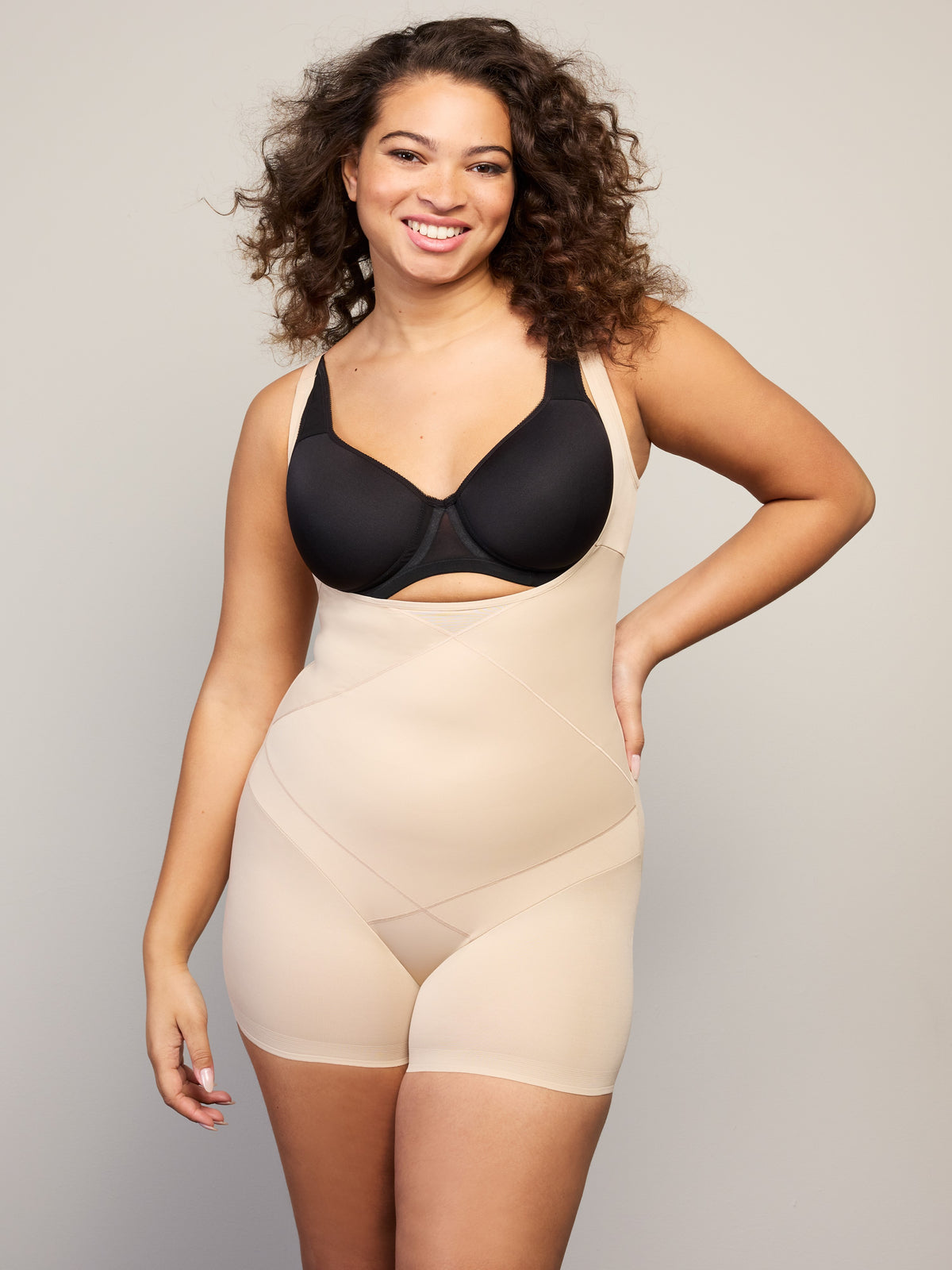 a woman wearing a black open bust tummy control bodysuit against a beige wall all-groups